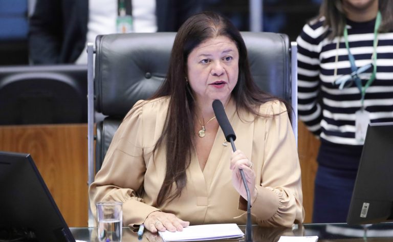 Deputada Laura Carneiro fala ao microfone. Ela é clara, tem cabelo escuro e comprido e veste uma blusa bege