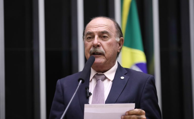 Deputado Dr. Zacharias Calil fala ao microfone, Ele é claro, tem cabelo escuto e bigode, usa um terno escuro e segura um papel