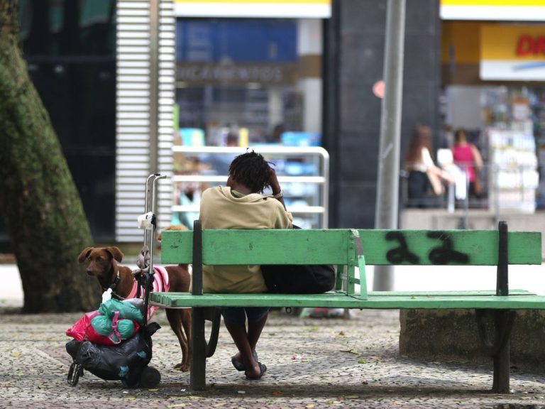 Pessoa sem teto está sentada no banco de uma praça com uma sacola e um cachorro ao lado