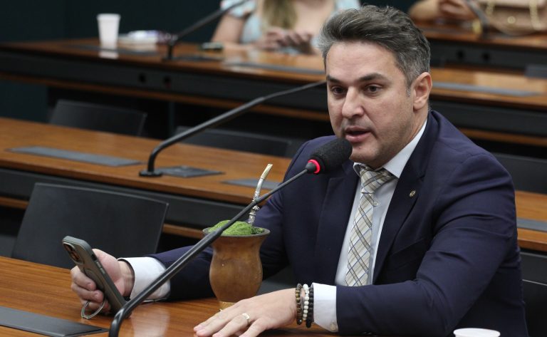 Deputado Zucco fala ao microfone. Ele é um homem claro, com cabelo curto e escuro. Veste um terno azul-marinho e segura um papel