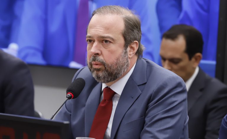 Um homem de bigode e barba fala ao microfone. Ele é branco e usa um terno cinza escuro com uma gravata vermelha