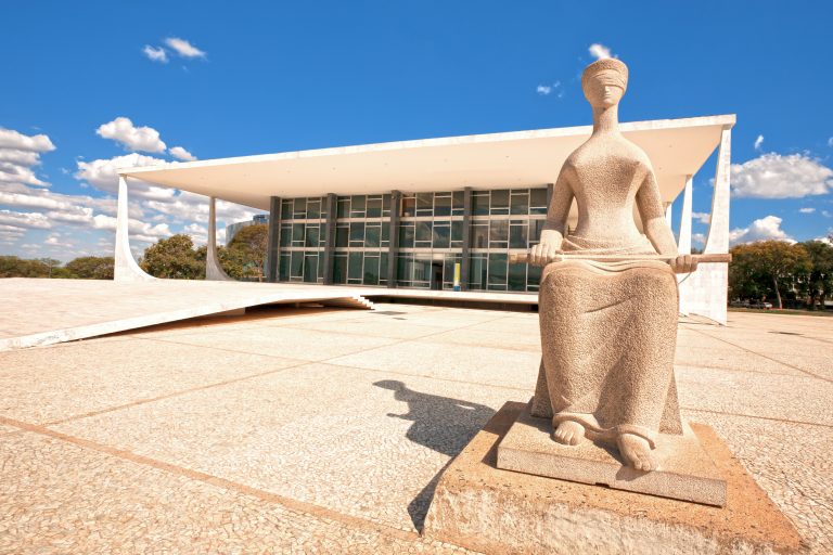 Fachada do STF: um prédio branco com vidraças ao fundo, em primeiro plano a estátua de uma mulher vendada sentada segurando um bastão