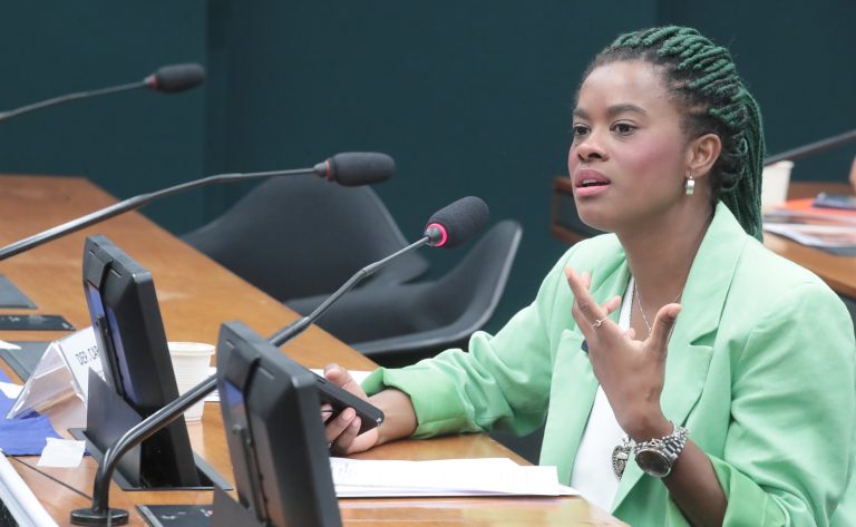 Deputada Carol Dartora fala ao microfone. Ela é negra, tem o cabelo verde trançado, usa um blazer verde e segura um celular