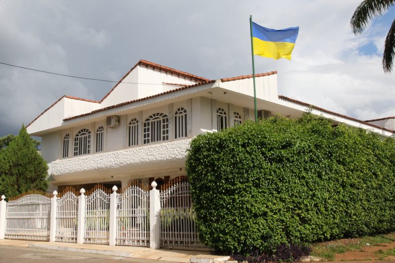 Fachada de uma casa de dois andares branca, com uma cerca viva na lateral e uma bandeira da Ucrânia hasrteada