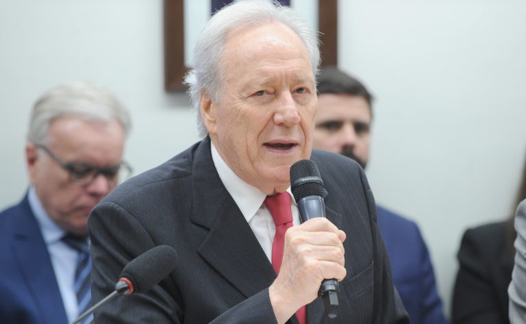 Um home claro com cabelos brancos, usando terno escuro, fala ao microfone