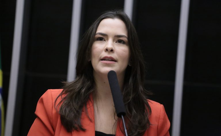 Deputada Maria Arraes fala ao microfone. Ela é clara, tem cabelos escuros, lisos e compridos; e usa um blazer cinza