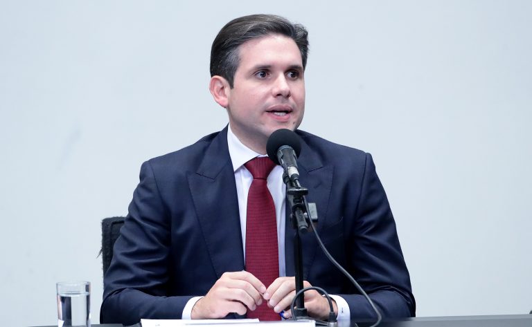 Hugo Motta fala ao microfone. Ele é um homem branco, tem o cabelo escuro, veste um terno azul-marinho com camisa branca e gravata vermelha