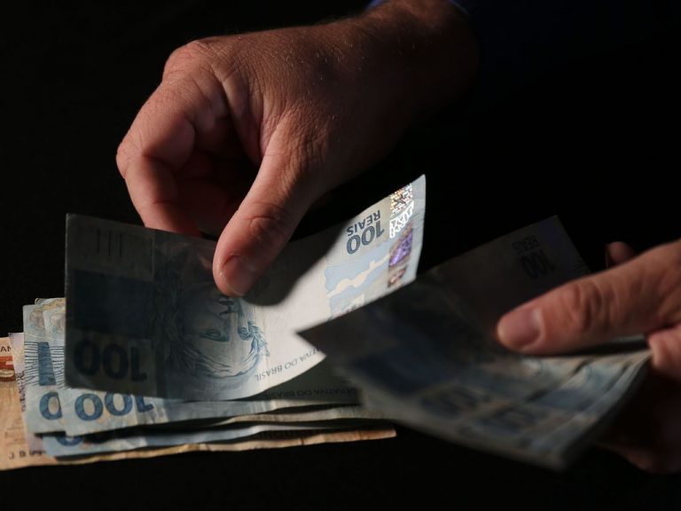 Imagem mostra mãos com dinheiro (como se alguém tivesse contando o dinheiro) e um fundo escuro