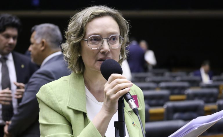Deputada Maria do Rosário fala ao microfone. Ela é uma mulher clara, com cabelo loiro, curto e olhos claros, usa óculos e veste um blazer verde claro